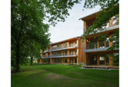 Blick aus den Bäumen auf dei Wohnbebauung in Holzbauweise und großzügigen Fenstertüröffnungen zu Balkonen und Terrassen