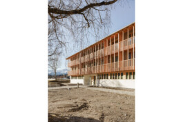 Schräge Hofansicht des kommunanlen Wohnungsbaus in Oberau. Erdgeschoss in MAssivbauweise sichtbar, ab dem Obergeschoss mit Holz verkleidet in vertikaler Struktur