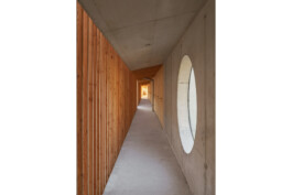 Blick in den Flur des Nebengebäudes mit Kellerersatzräumen. Rechts Sichtbetonwand mit rundem Fenster zur BAhnlinie, links vertikale Holzlattung als Raumtrenner der Kellerersatzräume