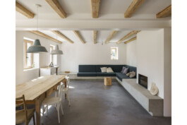 Blick RIchtung Wohnraum mit Sofa aus ORtbeton,gemauertem Holzofen, sichtbaren Deckenbalken und lasiertem Sichtestrich